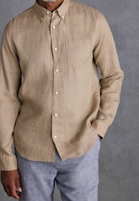 Beige linnen overhemd met lange mouwen, een button-down kraag, één borstzak en lichte knopen, casual gedragen met textuur lichtblauwe broek.