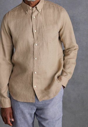 Chemise en lin beige à manches longues avec col à revers, poche poitrine unique et boutons clairs, portée de manière décontractée avec un pantalon bleu clair texturé.