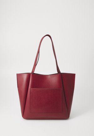 Sac cabas en cuir rouge avec de longues anses et une grande poche avant, sur un fond clair uni.