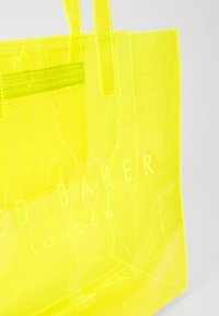Klare gelbe Tragetasche aus PVC mit zwei Trägern, ausgestattet mit einer Reißverschlusstasche und dem aufgedruckten Text "TED BAKER LONDON" auf der Vorderseite.