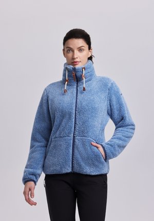 Mujer con chaqueta de forro polar azul claro con cuello alto y cordones, de pie con una mano en el bolsillo contra un fondo liso.