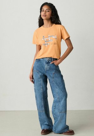 Orange cotton t-shirt with a graphic text design in black, paired with oversized blue denim jeans featuring side pockets and stitched details.