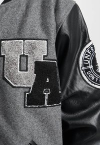 Grå ullblanding jakke med svarte skinnærmer, med hevet "UA" bokstaver og en rund lapp med hvite bokstaver på armen.