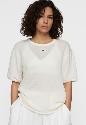 Top in maglia oversize color crema con maniche corte, scollatura rotonda e una piccola etichetta del logo sul petto. Tessuto testurizzato, vestibilità comoda.