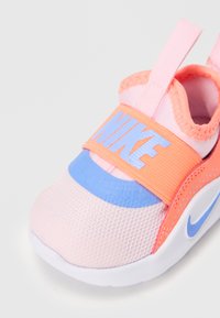 Otroški čevlji Nike z lahkotnim mrežastim zgornjim delom v roza in oranžni barvi, z modrim trakom in belim podplatom. Teksturne podrobnosti in logotip.
