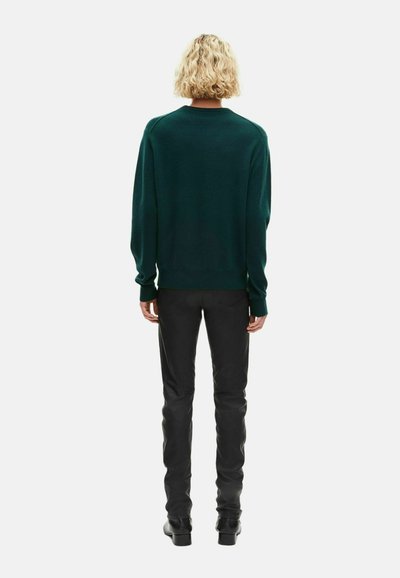 Osoba z krótkimi, kręconymi blond włosami, ubrana w ciemnozielony sweter, czarne spodnie i czarne buty, stojąca tyłem na jednolitym tle.