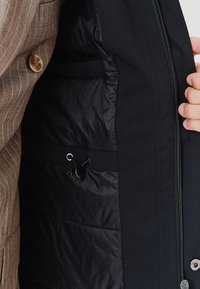 Wnętrze czarnej kurtki ma pikowaną strukturę, lewą kieszeń oraz sznurek ze metalowymi elementami. Zewnętrzna warstwa ma gładkie wykończenie.