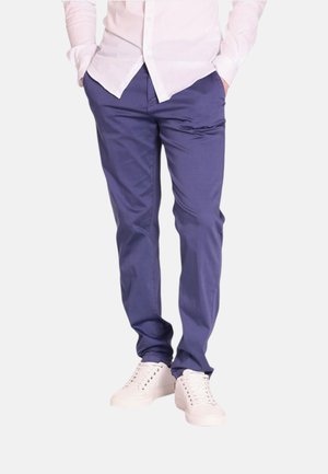 Uomo che indossa pantaloni slim fit blu, sneaker bianche e una camicia rosa chiaro a maniche lunghe con le mani in tasca.