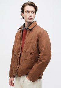 Jachetă din pânză maro cu guler din catifea, două buzunare frontale, închidere cu fermoar și detalii cu inel metalic, purtată deasupra unui pulover roșu.