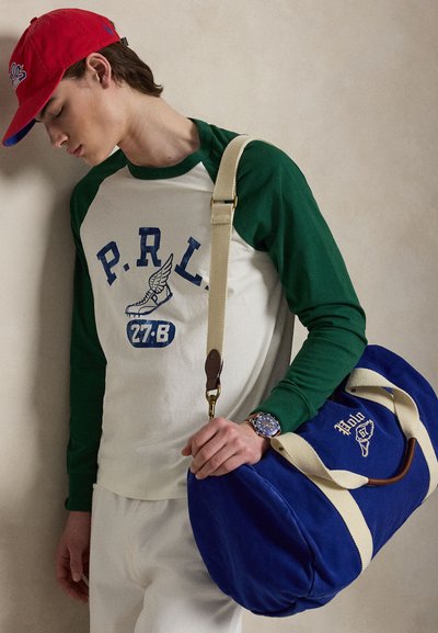 Νεαρός άνδρας που φορά πράσινο και λευκό πουλόβερ με μακριά μανίκια, κόκκινο καπέλο, λευκό παντελόνι, κρατώντας μπλε σακ βουαγιάζ με κρεμ λουριά στον ώμο.