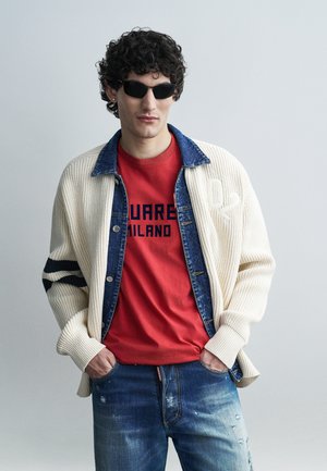 Férfi fekete napszemüvegben, piros mintás pólóban, farmerdzsekiben, krémszínű kötött kardigánban és kék kopott farmernadrágban, egyszínű háttér előtt.