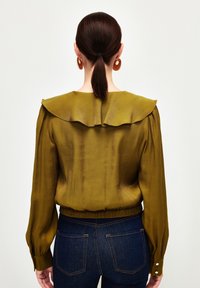 Femme à queue de cheval brune portant un chemisier à manches longues vert olive avec un col à volants et un jean en denim bleu foncé, vue de dos.