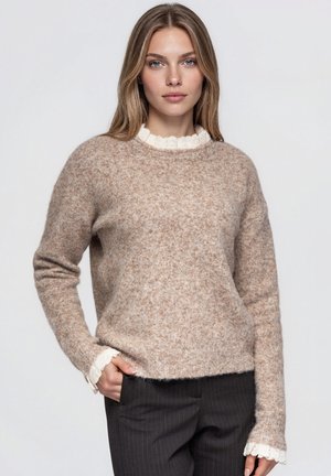 Modèle portant un pull beige texturé sur un chemisier blanc à volants et un pantalon sombre à fines rayures, devant un fond clair uni.
