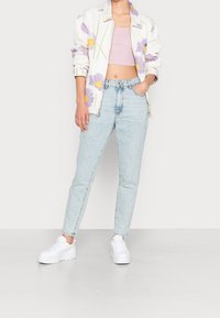 Person klädd i ljusblå högmidjade jeans, vit blommig jacka med lila blommor, ljusrosa crop top och vita platåskor, stående.