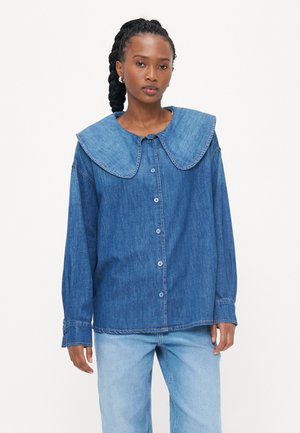 Denimskjorte med stor, rund krage, knappelukking foran og lange ermer. Stoffet er solid mørkeblått med synlige sømdetaljer.