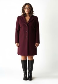 Abito blazer bordeaux con colletto a rever, bottoni frontali e due tasche laterali. Indossato con stivali alti in pelle nera. Texture liscia.