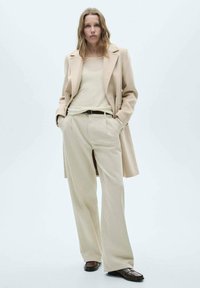Beige ullkappa över krämfärgad topp och ljusbeige vida byxor, accentuerad med ett brunt bälte och bruna loafers. Släta texturer, minimalistisk design.