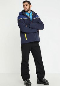 Giacca blu navy con dettagli gialli e cerniera, tessuto impermeabile, dotata di cappuccio; abbinata a pantaloni da sci neri e stivali da neve.