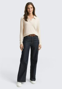 Beige Langarm-Polohemd mit Kragen, kombiniert mit dunkelblauen weit geschnittenen Jeans. Ein brauner Gürtel und weiße Sneakers runden das Outfit ab.