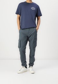 Granatowy t-shirt z okrągłym dekoltem i białym nadrukiem, zestawiony z ciemnoszarymi spodniami cargo z kieszeniami i mankietami. Białe trampki dopełniają stylizację.