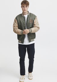 Giacca bomber verde e beige con un patch testurizzato a forma di "B", polsini a righe e chiusura frontale con bottoni, abbinata a una camicia bianca e pantaloni scuri.