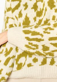 Sweter z dzianiny w kolorze kremowym z zielonym abstrakcyjnym wzorem w lamparta, z prążkowanymi mankietami i miękką, teksturowaną tkaniną. Zbliżenie na wzór rękawa.