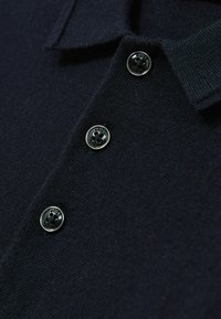 Polo majica tirkizne boje izrađena od pletenog materijala, s tri sjajna crna dugmeta na ovratniku za zatvaranje.