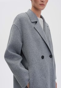 Grijze wolblendjas met een dubbelgebreid ontwerp, grote geknipte kraag en twee zwarte knopen, met relaxte mouwen en zijzakken.