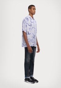Camisa corta de manga corta en tono púrpura claro con un patrón azul en espiral, combinada con jeans oscuros y zapatillas negras que presentan texto en blanco.