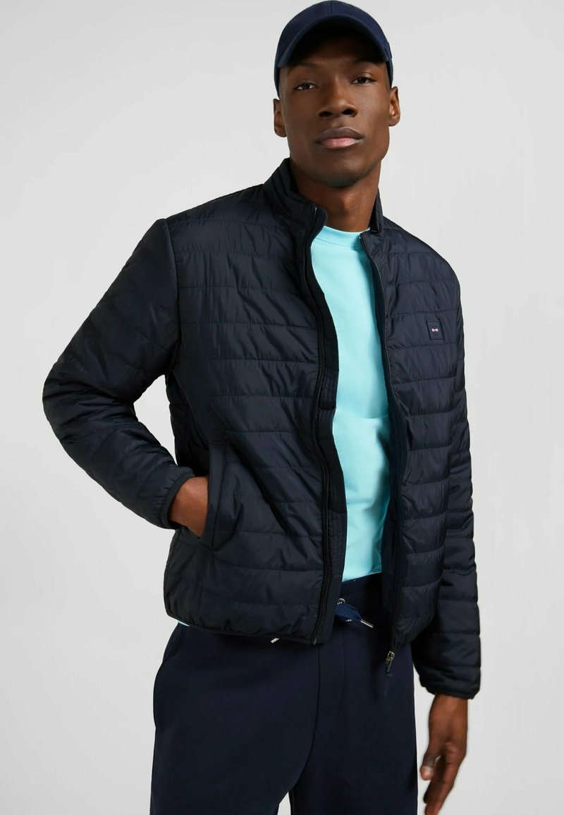 Veste matelassée marine avec fermeture éclair intégrale et col montant, associée à un t-shirt bleu clair à col rond. Le mannequin a les mains dans les poches et porte une casquette marine.