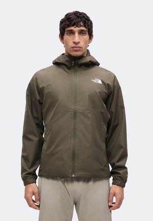 Mand iført en grøn North Face hættejakke med lynlås foran og lyse bukser mod en ensfarvet baggrund.