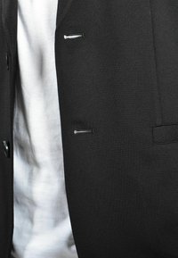 Giacca nera con una texture liscia, dotata di piccole tasche frontali e chiusure a bottoni. Indossata sopra una camicia bianca.