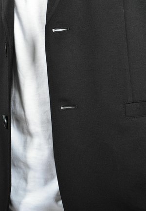 Giacca nera con una texture liscia, dotata di piccole tasche frontali e chiusure a bottoni. Indossata sopra una camicia bianca.