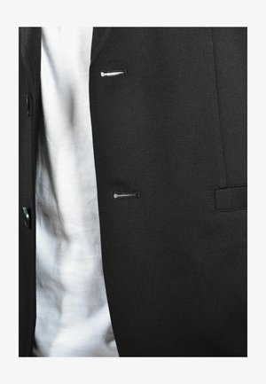 Giacca nera con una texture liscia, dotata di piccole tasche frontali e chiusure a bottoni. Indossata sopra una camicia bianca.