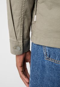 Jachetă verde deschis, cu nasturi, cu o textură netedă, având manșete cu închidere cu buton, asociată cu blugi din denim albastru.