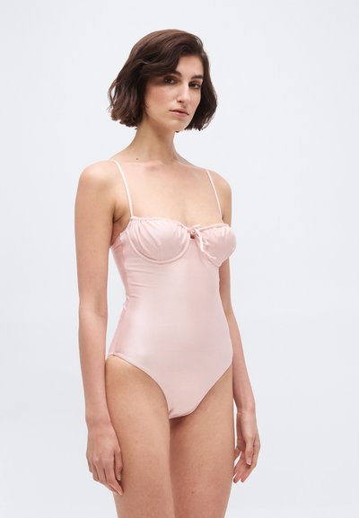 Kobieta o krótkich ciemnych włosach, ubrana w jasnoróżowy jednoczęściowy strój kąpielowy z cienkimi ramiączkami i małą kokardką z przodu, stojąca na tle jednolitej białej ściany.