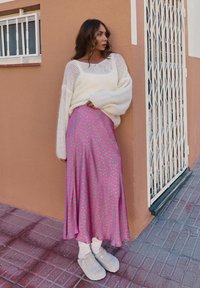 Woman wearing a white knit sweater, pink patterned skirt, white socks, and beige clogs leaning against a peach-colored wall near a white metal gate.