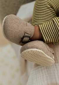 Chaussons en feutre doux de couleur beige clair, à la forme arrondie, avec un détail de boucle sécurisé et des semelles texturées blanches.