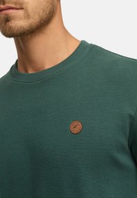 Mann trägt ein geripptes, dunkelgrünes Rundhalsausschnitt-Shirt mit kleinem, rundem braunem Leder-Logo-Patch auf der Brust vor einem schlichten Hintergrund.