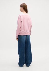 Felpa rosa con scollatura arrotondata e polsini a costine abbinata a pantaloni in denim larghi di un blu scuro. Texture liscia, vestibilità rilassata, dettagli minimi.