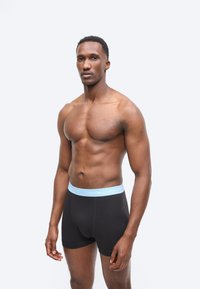 Muskulárny muž stojaci, oblečený v čiernych boxerkách s pásom Calvin Klein svetlomodrej farby, na jednoduchom svetlosivom pozadí.