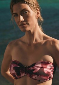 Top bikini bandeau in un motivo astratto rosa e bordeaux, con dettaglio a torsione al centro e accenti in hardware metallico.