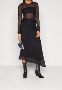 Top de encaje negro de mangas largas superpuesto sobre una falda maxí de encaje negro. Combinado con botas de vaquero negras con bordados blancos y un bolso plateado.