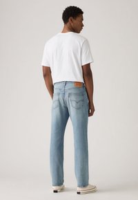 Calças de ganga azul claro com perna reta, apresentando um remendo de couro castanho na cintura e dois bolsos traseiros, usadas com uma t-shirt branca.