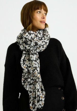 Femme portant un pull noir et une écharpe noire et blanche à fleurs, regardant sur le côté contre un fond clair uni.