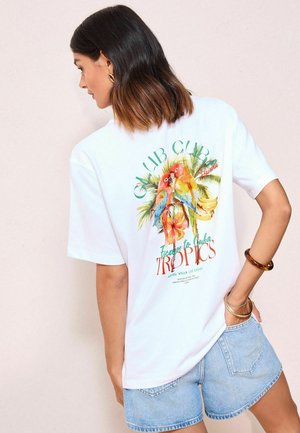 T-shirt en coton blanc avec un imprimé tropical coloré comprenant deux perroquets et du texte, associé à un short en denim bleu clair.