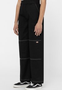Pantalon large noir avec coutures contrastantes blanches, en tissu durable. Comprend un patch logo sur le côté et un design taille haute.