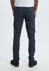 Navy dress pants made of textured fabric, feature two back pockets with buttons, and a tailored fit. Black shoes are worn with the pants.