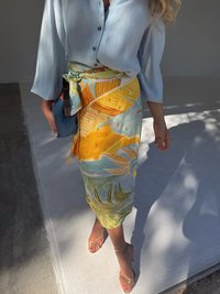 A woman in a light blue blouse holds a clutch, wearing a vibrant yellow skirt with tropical patterns and strappy clear heels.