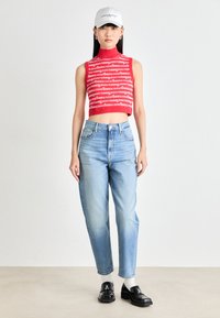 Top court rouge avec motif de texte répété, jean taille haute bleu clair, mocassins noirs et une casquette de baseball blanche. Fond de surface plate.